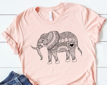 May include: A pink t-shirt with a black and white mandala elephant design. The elephant has a small black heart on its side. The shirt is paired with blue jeans and white sneakers.