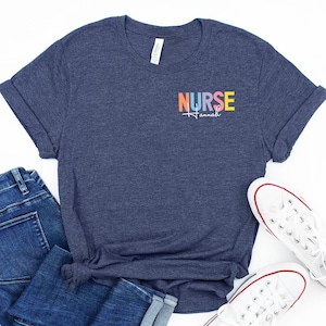 May include: A navy blue t-shirt with the word "NURSE" in rainbow colors and a stethoscope graphic. The name "Hannah" is printed below the word "NURSE". The shirt is paired with blue jeans and white sneakers.