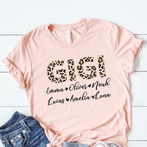 May include: Pink t-shirt with the word "GIGI" in leopard print letters. Below the word are the names "Emma, Oliver, Noah, Lucas, Amelia, Luna" with hearts between each name.