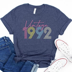 May include: A navy blue t-shirt with the text "Vintage 1992" in a retro font with colorful stripes. The shirt is paired with blue jeans and white sneakers.