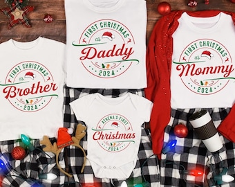 May include: A set of four white t-shirts with red and green Christmas-themed designs. The shirts say "First Christmas as Daddy", "First Christmas as Mommy", "First Christmas as Brother", and "Athena's First Christmas".