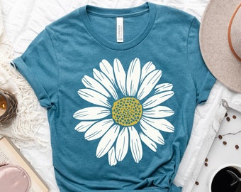 May include: Teal t-shirt featuring a large white daisy with a yellow center. The shirt is displayed on a white surface with a hat, coffee, and a book. The shirt is made of a soft material.