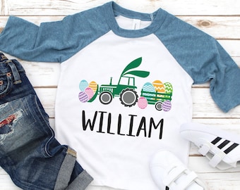 May include: White and blue raglan style shirt with a green tractor graphic carrying Easter eggs and the name "WILLIAM". The shirt is paired with blue jeans and white sneakers with black stripes.