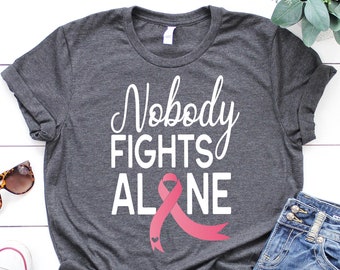 Op de afbeelding: Een grijs T-shirt met de tekst "Nobody Fights Alone" in witte letters en een roze lint met een klein hartje.