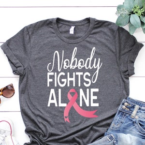 May include: A gray t-shirt with the text "Nobody Fights Alone" in white lettering and a pink ribbon with a small heart design.