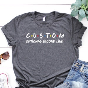 May include: A dark gray t-shirt with a white graphic that says "C.U.S.T.O.M" with colorful dots and "OPTIONAL SECOND LINE" underneath. The shirt is laid out on a white wooden surface with a pair of sunglasses, a pair of white sneakers, and a pair of blue jeans.