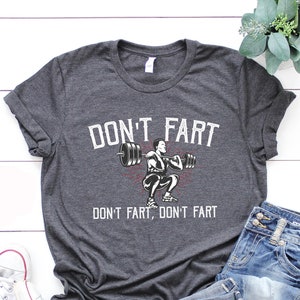 May include: A dark heather grey t-shirt with a white graphic of a person lifting weights. The text "DON'T FART, DON'T FART" is printed on the shirt.
