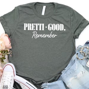 May include: A casual flat lay featuring a gray t-shirt with the text "PRETTI-GOOD. Remember" in white, paired with light-wash denim jeans, white sneakers, and a coffee cup. Soft pink flowers and a bracelet complete the ensemble.