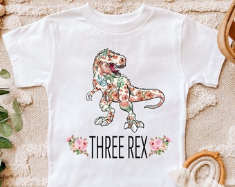 Può includere: Maglietta bianca con un dinosauro stampato floreale e il testo "THREE REX" con accenti floreali.