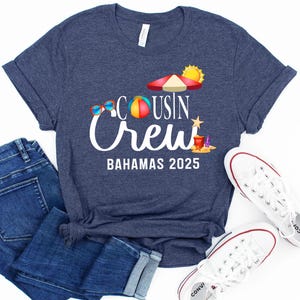 May include: Navy blue t-shirt with the text "Cousin Crew Bahamas 2025" and beach-themed graphics. The shirt is paired with blue jeans and white sneakers. A fun and casual outfit for a beach vacation.