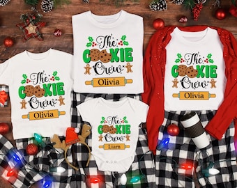 May include: White t-shirts and a onesie with the text "The Cookie Crew" in green and red, featuring cookie and gingerbread man graphics. The shirts are paired with black and white plaid pajama pants. Festive holiday decor surrounds the clothing.