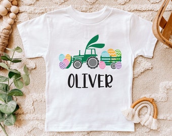 May include: White toddler t-shirt featuring a green tractor pulling a wagon filled with Easter eggs. The tractor has bunny ears. The name "OLIVER" is printed in black below the graphic. The shirt is on a neutral background.