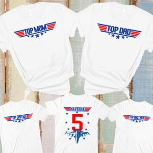 May include: White t-shirts with Top Gun-inspired designs. The shirts feature text like "Top Mom," "Top Dad," "Top Sister," and "Top Brother," along with a "Maverick" shirt with the number 5 and a jet graphic. Each design has red, white, and blue accents.