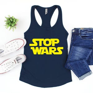 May include: A navy blue racerback tank top with the words "STOP WARS" in yellow block letters. The tank top is paired with blue jeans and white sneakers with red accents. A small potted plant is also in the image.