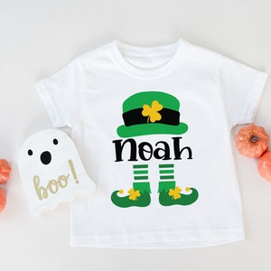 May include: White t-shirt with a green leprechaun graphic and the name "Noah" printed on it. The leprechaun is wearing a green hat with a gold buckle and has a green beard.