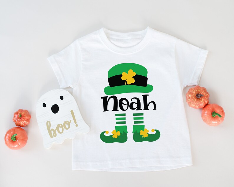 May include: White t-shirt with a green leprechaun graphic and the name "Noah" printed on it. The leprechaun is wearing a green hat with a gold buckle and has a green beard.
