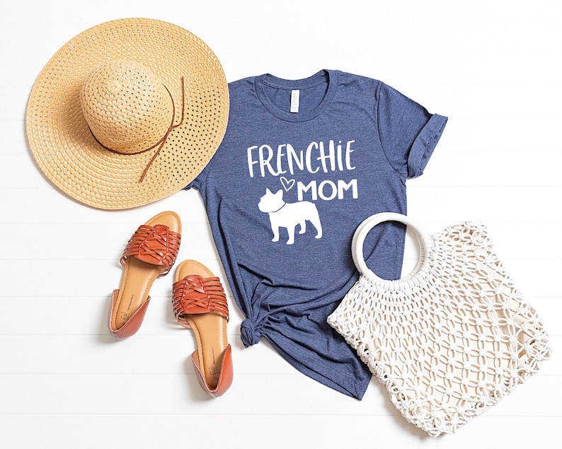 May include: A navy blue t-shirt with the text "Frenchie Mom" and a white silhouette of a French Bulldog. The shirt is laid out on a white surface with a straw hat, brown sandals, and a white crochet bag.