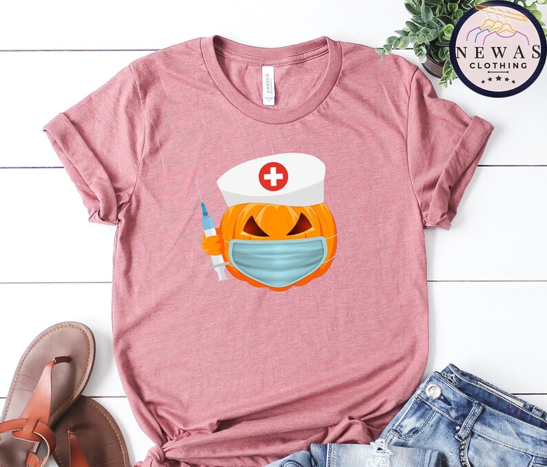 May include: A pink t-shirt with a graphic of a pumpkin wearing a nurse's hat and a face mask. The pumpkin is holding a syringe.