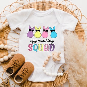 May include: White baby onesie with a graphic of four pastel-colored Easter bunnies wearing sunglasses. The text "egg hunting SQUAD" is printed below the bunnies.