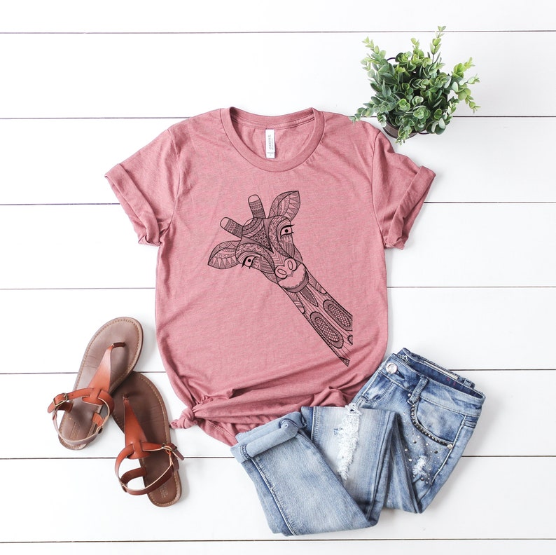 May include: A pink t-shirt with a black and white patterned giraffe design. The giraffe's head is peeking out from the left side of the shirt.