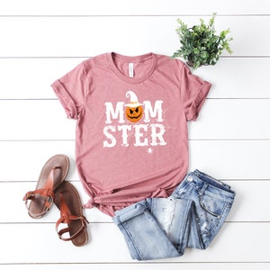 May include: A mauve t-shirt with the text "MOMSTER" in white with a pumpkin wearing a witch hat in the middle of the word. The pumpkin has a scary face and a white witch hat. There is a spider web behind the pumpkin and a small spider below the word.