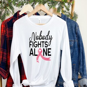 May include: White long-sleeve t-shirt with the text "Nobody Fights Alone" and a pink ribbon graphic.