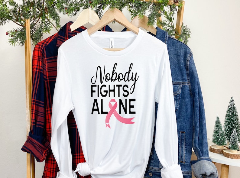 May include: White long-sleeve t-shirt with the text "Nobody Fights Alone" and a pink ribbon graphic.