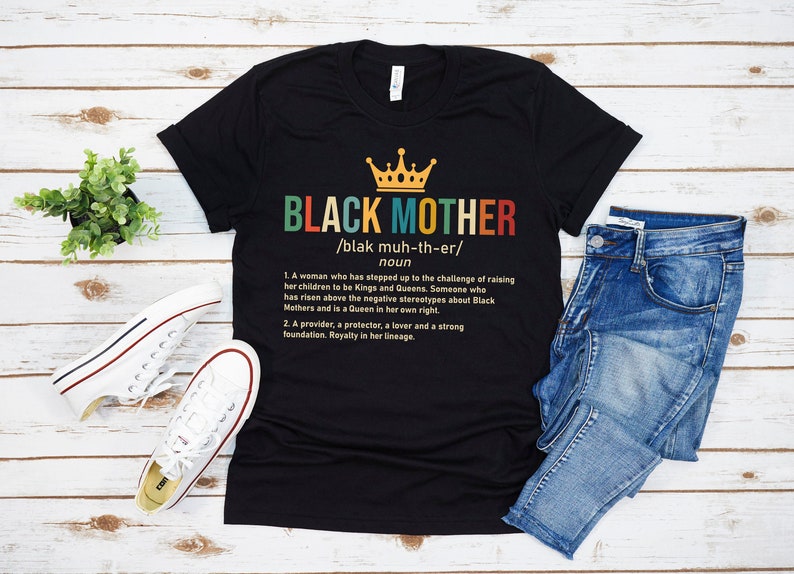 May include: Black t-shirt with a crown and the words "BLACK MOTHER" defined as a noun. The definition reads: "A woman who has stepped up to the challenge of raising her children to be Kings and Queens. Someone who has risen above the negative stereotypes about Black Mothers and is a Queen in her own right. A provider, a protector, a lover and a strong foundation. Royalty in her lineage."