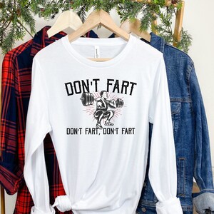 K&ouml;nnte beinhalten: Wei&szlig;es Langarmshirt mit einer schwarzen Grafik einer Person, die Gewichte hebt. Der Text "Don't fart, don't fart" ist auf das Shirt gedruckt.