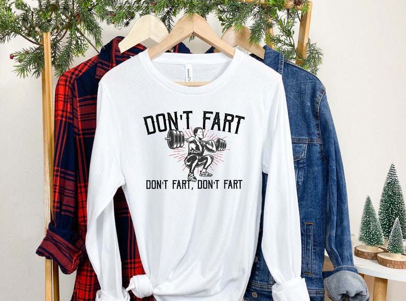 K&ouml;nnte beinhalten: Wei&szlig;es Langarmshirt mit einer schwarzen Grafik einer Person, die Gewichte hebt. Der Text "Don't fart, don't fart" ist auf das Shirt gedruckt.