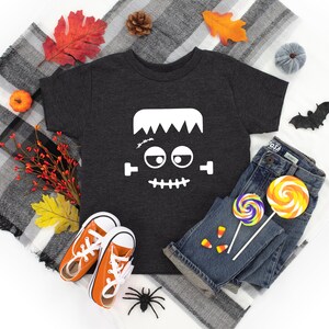 May include: A dark gray t-shirt with a white Frankenstein face graphic. The shirt is laid out on a white surface with a plaid blanket, fall leaves, a small pumpkin, a spider, and candy.