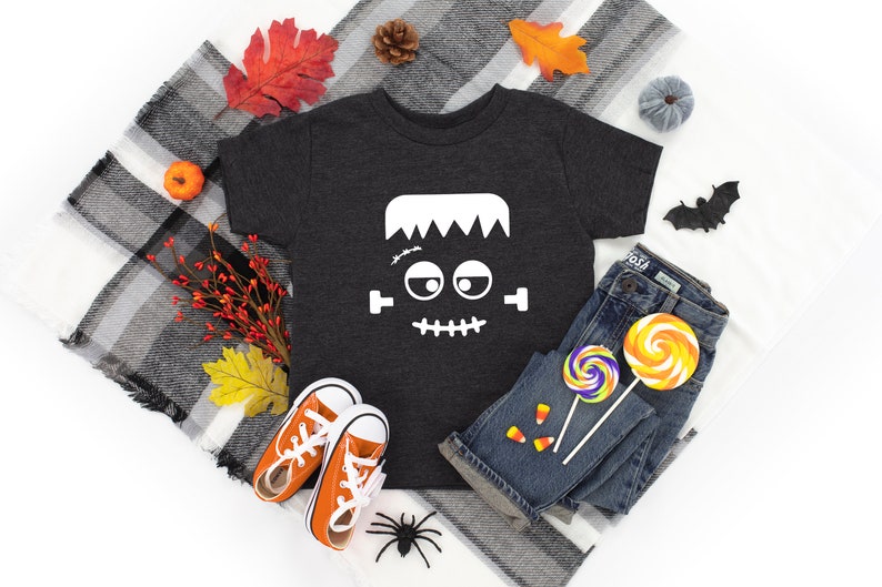 May include: A dark gray t-shirt with a white Frankenstein face graphic. The shirt is laid out on a white surface with a plaid blanket, fall leaves, a small pumpkin, a spider, and candy.