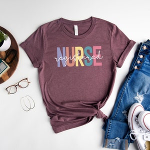 Pu&ograve; includere: Una maglietta color marrone rossiccio con la scritta "NURSE registered" in colori arcobaleno. La maglietta &egrave; abbinata a jeans blu e scarpe da ginnastica bianche.