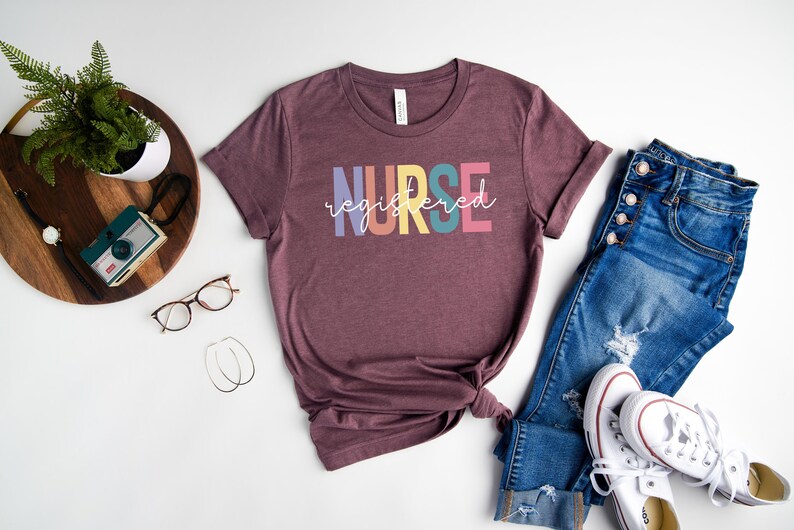 Pu&ograve; includere: Una maglietta color marrone rossiccio con la scritta "NURSE registered" in colori arcobaleno. La maglietta &egrave; abbinata a jeans blu e scarpe da ginnastica bianche.