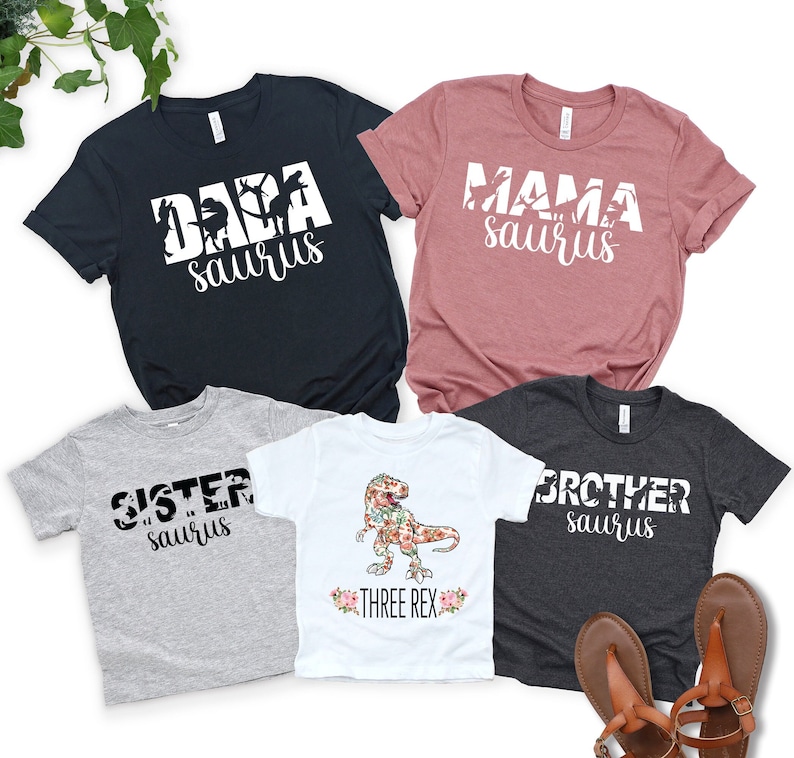 May include: A set of four t-shirts with a dinosaur theme. The shirts say "Dada saurus", "Mama saurus", "Sister saurus", and "Brother saurus". One shirt features a floral dinosaur with the text "Three Rex".