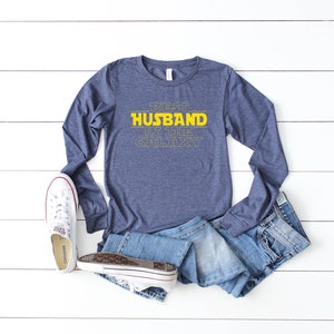 May include: A navy blue long-sleeve shirt with the text "BEST HUSBAND IN THE GALAXY" in yellow. The shirt is paired with blue jeans and white sneakers with red accents. The items are arranged on a white wooden surface.