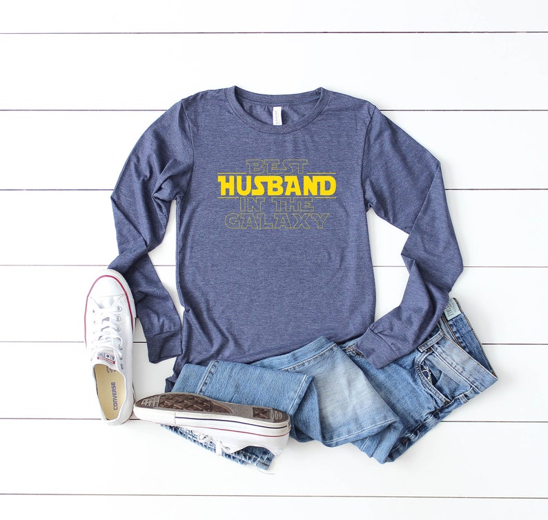 May include: A navy blue long-sleeve shirt with the text "BEST HUSBAND IN THE GALAXY" in yellow. The shirt is paired with blue jeans and white sneakers with red accents. The items are arranged on a white wooden surface.