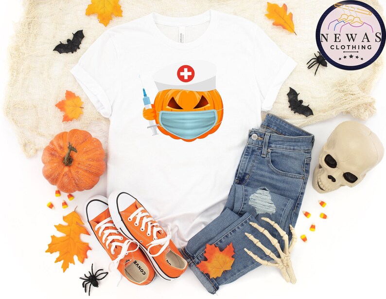 May include: White t-shirt with a pumpkin wearing a nurse's hat and a face mask. The pumpkin is holding a syringe. The shirt is laying on a white surface with a pair of orange converse shoes, a pair of blue jeans, a fake skull, and fall leaves.