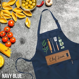 May include: Navy blue apron with a pocket and a wooden cutting board that says "Chef Liam". The apron has a design of kitchen tools, olive branches, and black olives.