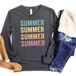 Puede incluir: Una camiseta de manga larga gris oscuro con la palabra "SUMMER" repetida cuatro veces en una pila vertical. Cada palabra es de un color diferente: verde azulado, amarillo, naranja y rosa. La camiseta est&aacute; colocada sobre un fondo blanco con un par de pisos tejidos marrones, un par de jeans azules y una boina de punto blanca.
