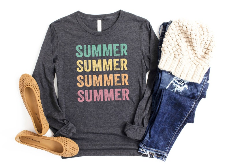 Puede incluir: Una camiseta de manga larga gris oscuro con la palabra "SUMMER" repetida cuatro veces en una pila vertical. Cada palabra es de un color diferente: verde azulado, amarillo, naranja y rosa. La camiseta est&aacute; colocada sobre un fondo blanco con un par de pisos tejidos marrones, un par de jeans azules y una boina de punto blanca.