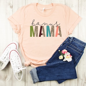 May include: A peach-colored t-shirt with the text "bonus mama" in colorful letters with a leopard print pattern. The shirt is paired with blue jeans and white sneakers.