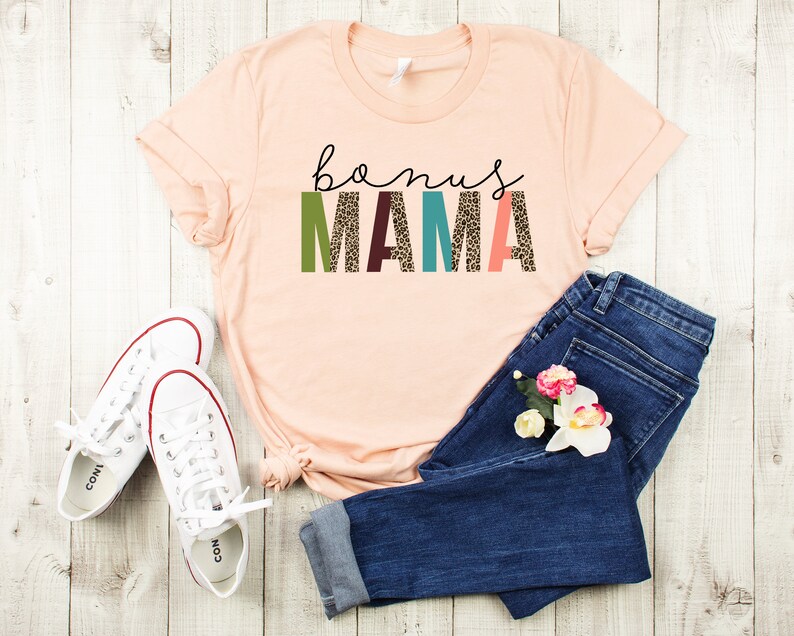 May include: A peach-colored t-shirt with the text "bonus mama" in colorful letters with a leopard print pattern. The shirt is paired with blue jeans and white sneakers.