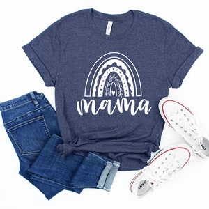 Pu&ograve; includere: Una maglietta blu navy con un design arcobaleno bianco e la parola "mamma" sotto. La maglietta &egrave; abbinata a jeans blu e scarpe da ginnastica bianche.