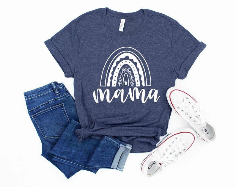 Pu&ograve; includere: Una maglietta blu navy con un design arcobaleno bianco e la parola "mamma" sotto. La maglietta &egrave; abbinata a jeans blu e scarpe da ginnastica bianche.