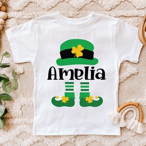 May include: White t-shirt with a green leprechaun design. The leprechaun is wearing a green hat with a black band and a gold buckle. The leprechaun's name is Amelia.