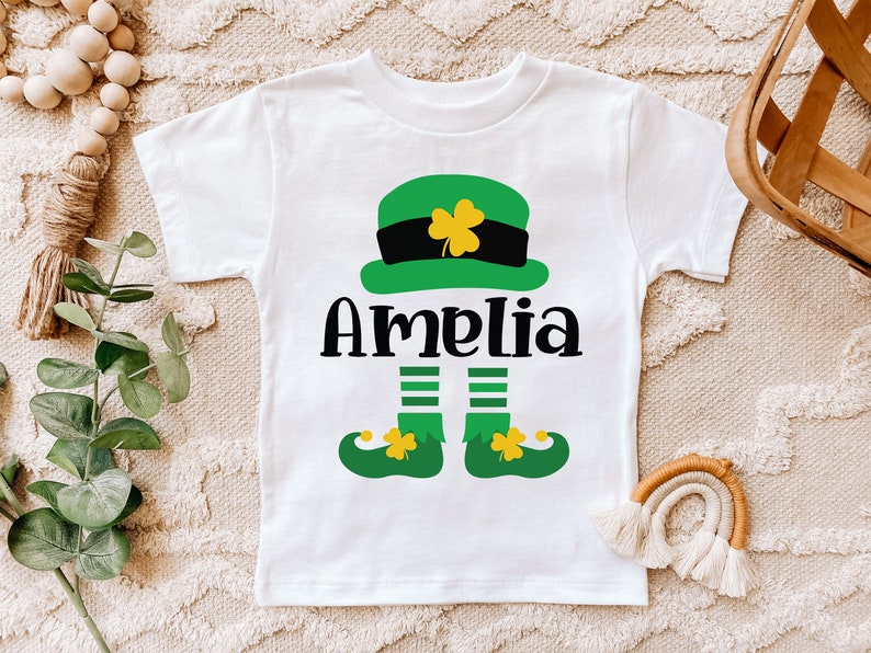 May include: White t-shirt with a green leprechaun design. The leprechaun is wearing a green hat with a black band and a gold buckle. The leprechaun's name is Amelia.