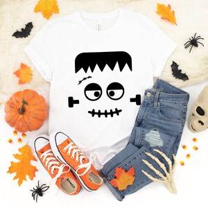 May include: White t-shirt with a black Frankenstein face graphic. The shirt is laid out on a white background with a pair of orange converse shoes, a pair of blue jeans, a pumpkin, a skull, and fall leaves.