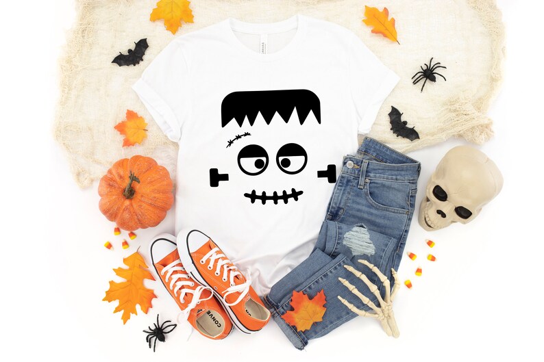 May include: White t-shirt with a black Frankenstein face graphic. The shirt is laid out on a white background with a pair of orange converse shoes, a pair of blue jeans, a pumpkin, a skull, and fall leaves.