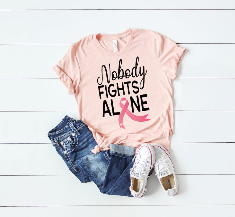 May include: Pink t-shirt with the text "Nobody Fights Alone" and a pink ribbon graphic. The shirt is laying on a white wooden surface with a pair of blue jeans and white sneakers.